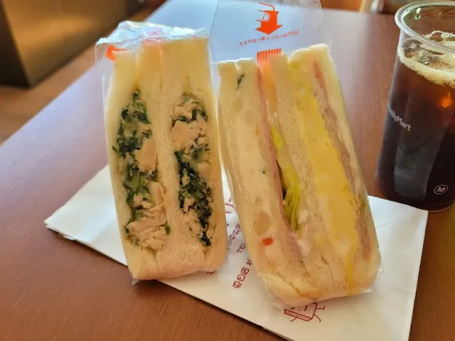 Two plastic wrapped sandwiches, one with chicken and greens and one with egg, beside a cold cup
