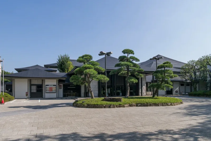 A low theatre building with a dark tiled roof, a paved forecourt and clipped pines