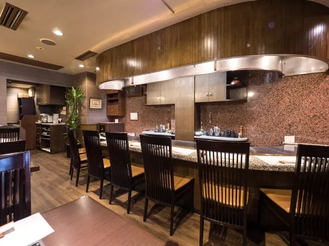 A long counter with a metal griddle, dark wooden chairs and a large hood above it
