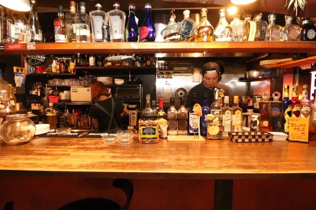 A wooden bar counter with bottles on high shelves and two staff behind it