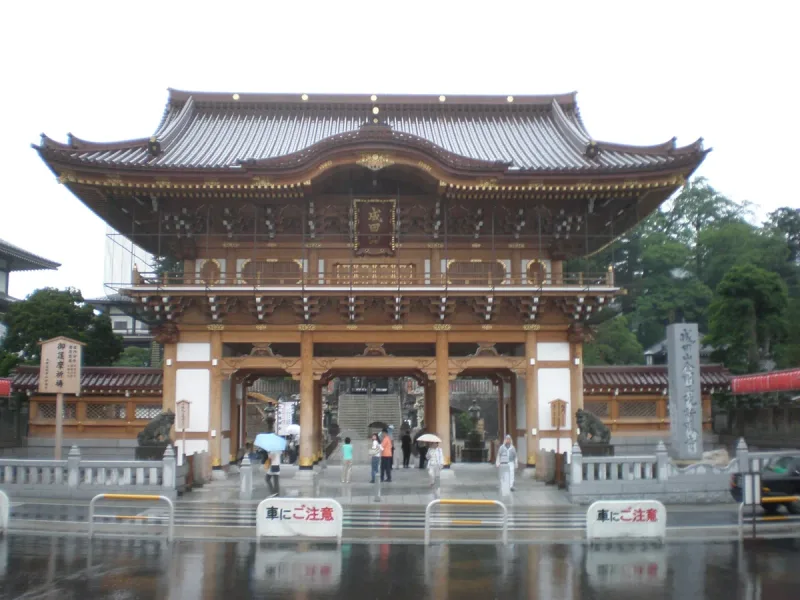 A large wooden temple gate in brown and gold, with visitors on the paved plaza in front