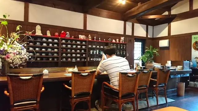 A wooden counter with a seated customer, and behind him shelves full of cups and small figurines