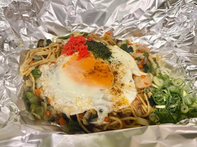 Fried noodles with vegetables and a fried egg on foil, scattered with green onion