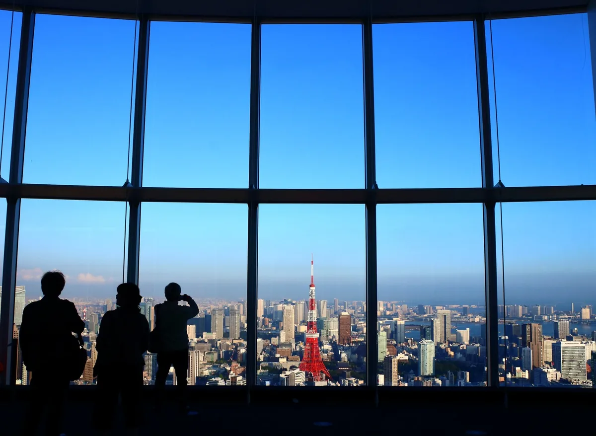 Silhouettes of visitors at a huge glass window looking out to a red and white tower and a city skyline