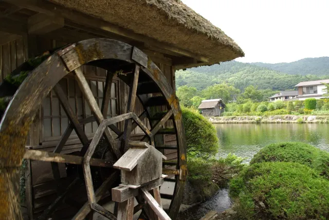 A wooden water wheel beside a clear pond and greenery