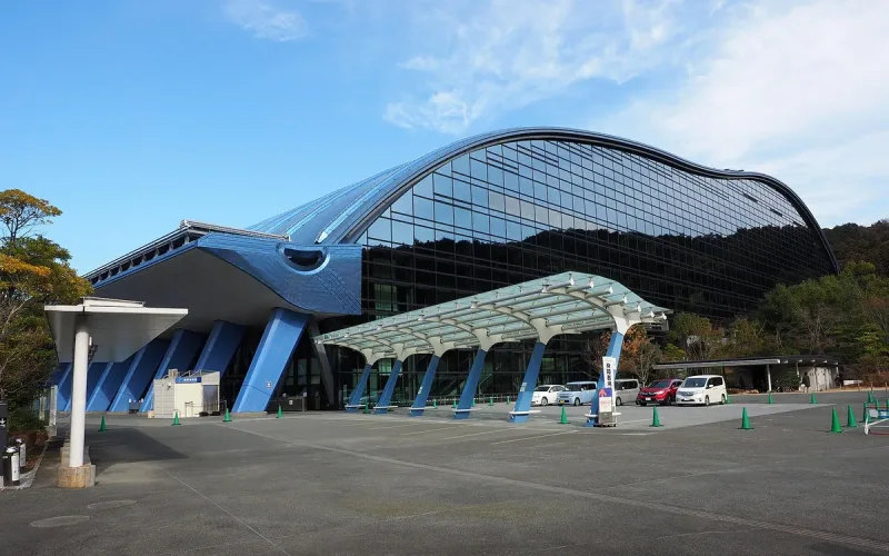 A large museum building with a curved blue roof and glass walls, against wooded hills