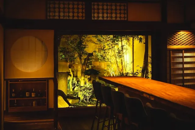 A wooden counter with stools facing a lit garden with trees and a stone lantern