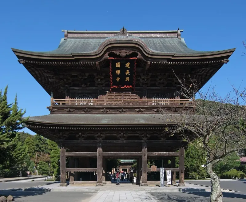 A two storey wooden temple gate with a broad tiled roof and visitors at the entrance below