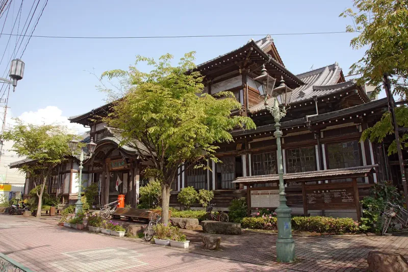 A historic wooden building with a heavy tiled roof, lanterns at the entrance and trees in front