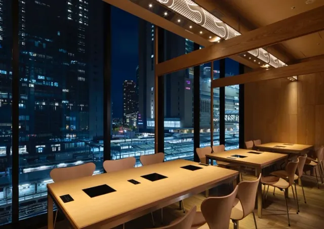 Long wooden tables facing a huge window, with the city buildings at night behind it