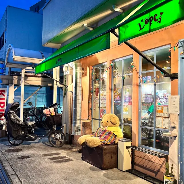 A facade lit at night with a green awning, colored bulbs and a bear doll on a crate