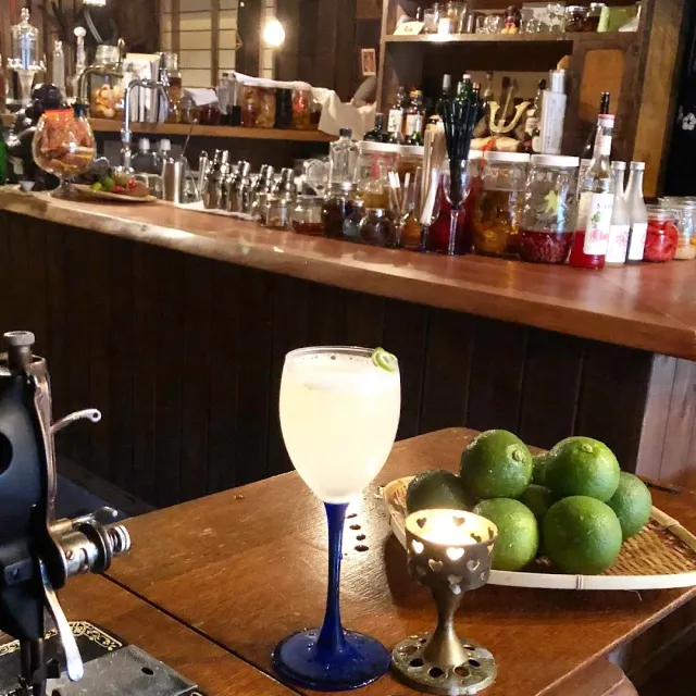 A cocktail glass with a blue stem beside green limes and a candle, facing a counter of jars