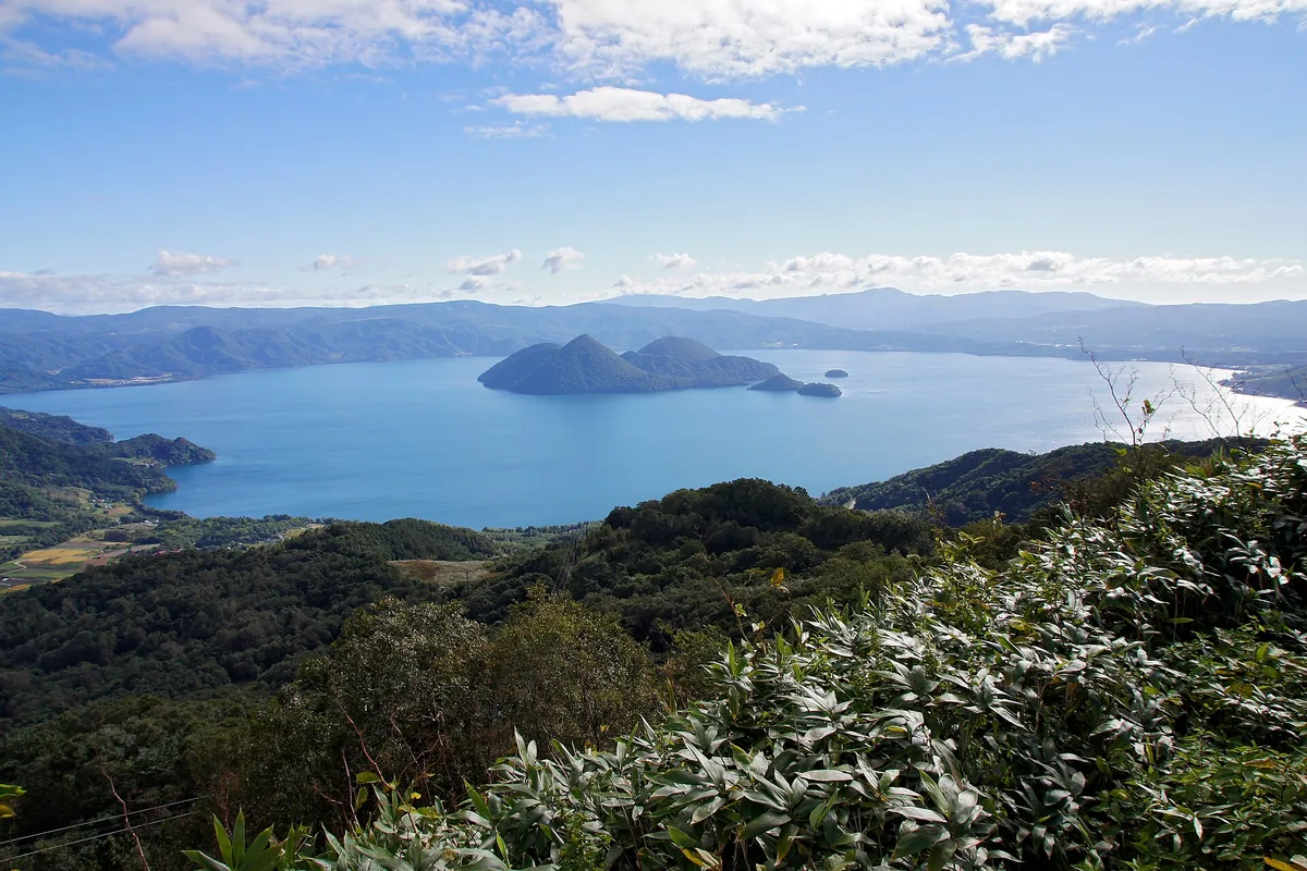 A wide blue lake with wooded islands at its centre, ringed by green hills under a bright sky
