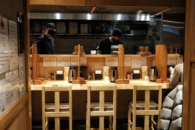 A pale wooden counter with stools, and behind it two staff in black in an open kitchen