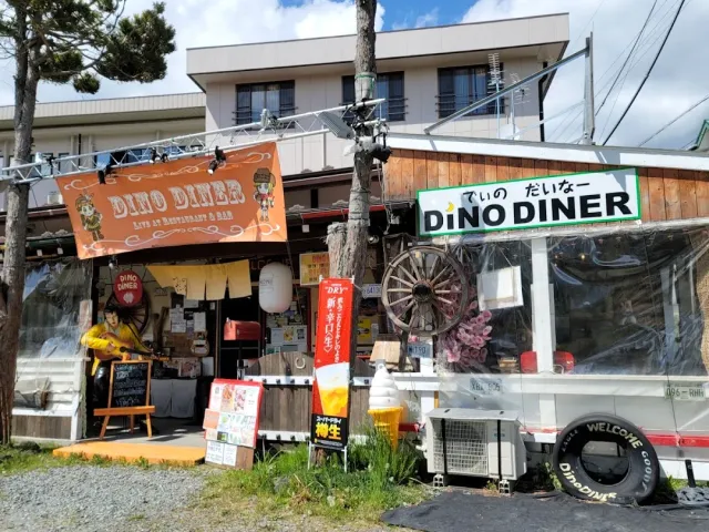 A low building with an orange banner, a green sign, a white trailer and an old cart wheel on the wall
