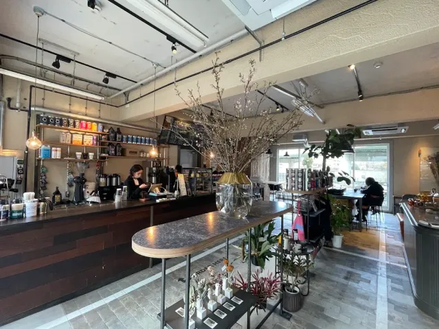 A space with a long counter, shelves of wares, a tall vase of branches and a worker behind the counter