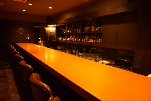 A long glossy counter with leather chairs facing bottle shelves in dim lighting