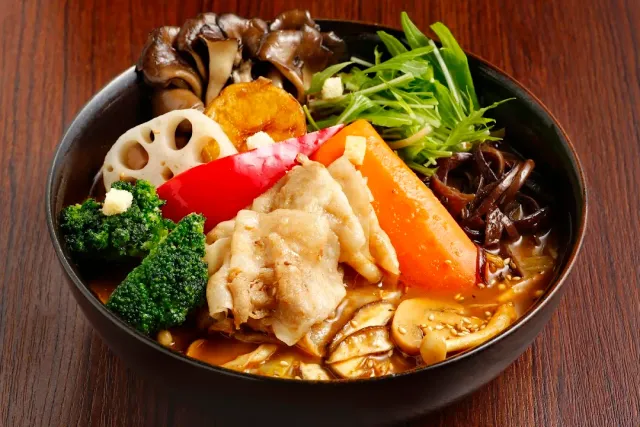 A dark bowl with brown soup, broccoli, carrot, lotus root, mushrooms and slices of meat