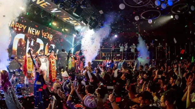 A dense crowd raising hands in front of a lit stage, with confetti and smoke in the air