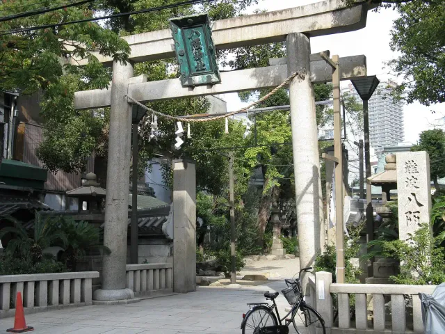 A stone torii gate at the entrance, with tall trees and buildings around