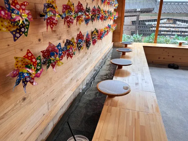 A wooden counter above a water channel, with seating mats and colorful paper pinwheels on the wall