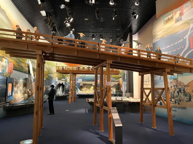 A full size wooden bridge inside an exhibition hall, with figures and displays beneath it