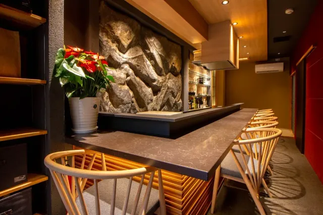 A long stone counter with light wooden chairs, a lit relief wall and a bunch of red flowers