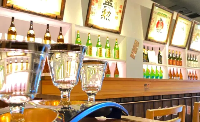 Glasses on a counter, and behind them bottle shelves and framed labels on the wall