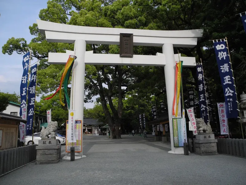 A white torii gate with long banners in front of a Shinto shrine, in a courtyard with trees