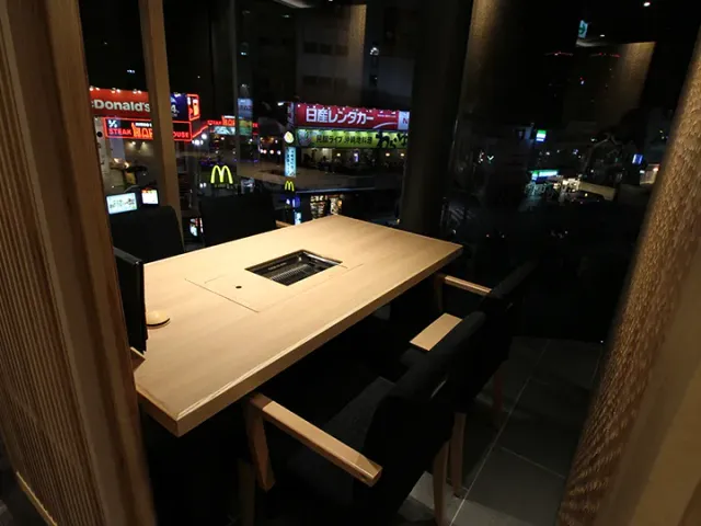 A private booth with a wooden table and a sunken grill, facing a window onto a neon filled street at night