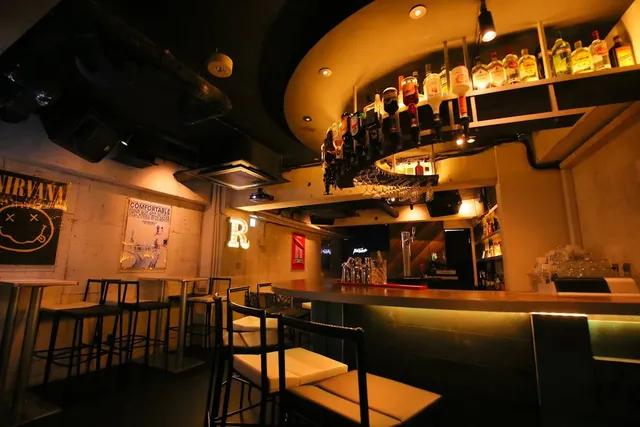 A dim bar with high chairs, bottles hanging above the counter and posters on a concrete wall