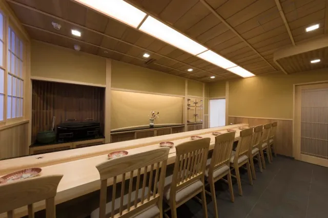 A room with a long pale wooden counter, a row of chairs and a lit wooden ceiling