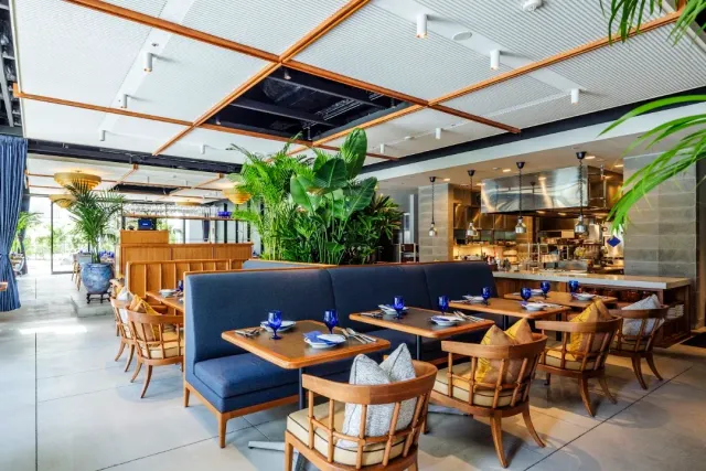 A wide hall with blue benches, wooden armchairs, tropical plants and an open kitchen