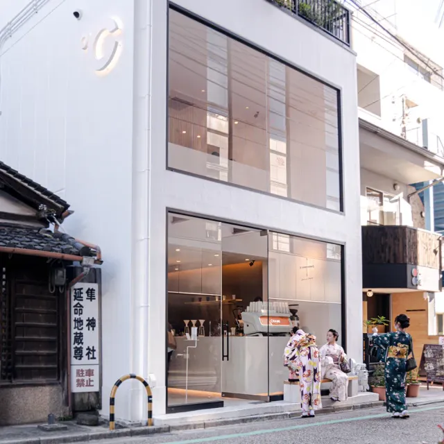 A white corner building with a glass front, with women in kimono sitting on a bench in front
