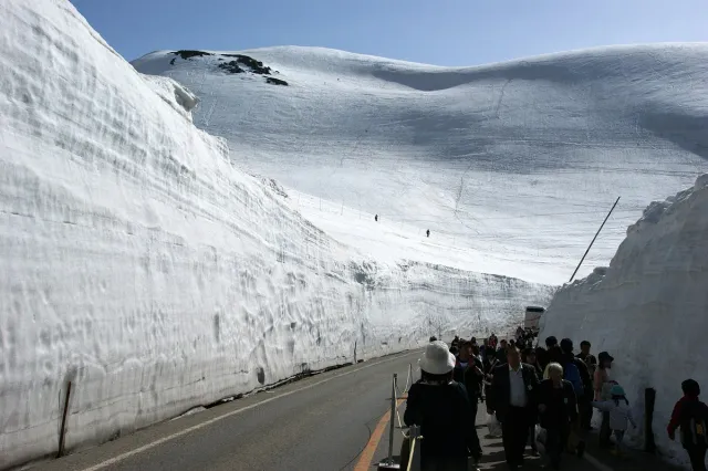A road cut between two towering walls of snow, with walkers