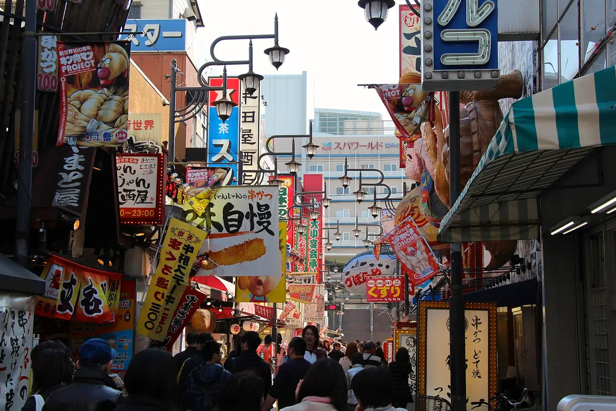 A crowded Shinsekai alley by day, packed with colourful kushikatsu signs and Billiken figures, with pedestrians filling the lane