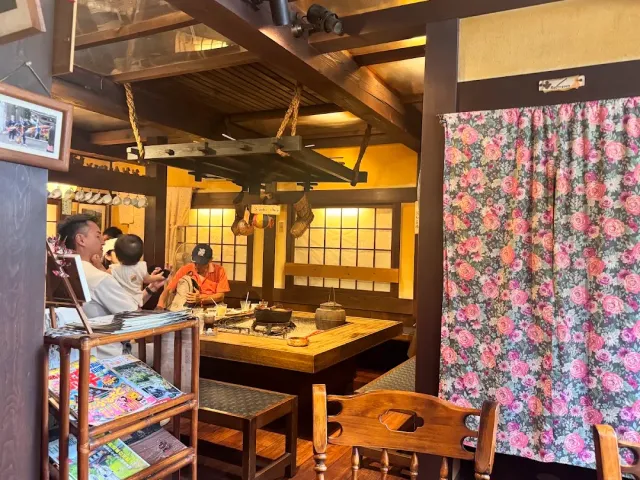 A wooden room with a hearth sunk in the table, a hanging beam, seated guests and a floral curtain