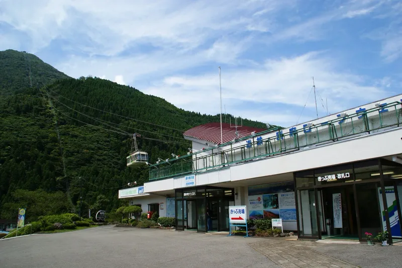 A ropeway station building on a wooded mountain slope, with a cabin hanging from the cable above