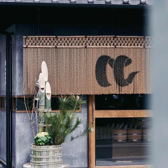 A string curtain with a black emblem above a dark front, beside a bamboo and pine branch decoration