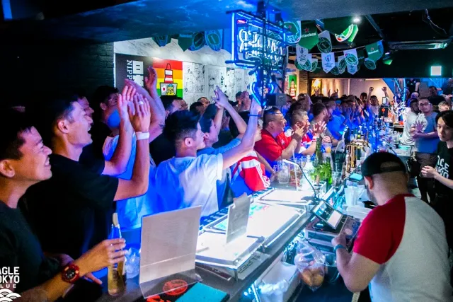 A dense crowd cheering along a bar counter in blue light with hanging pennants