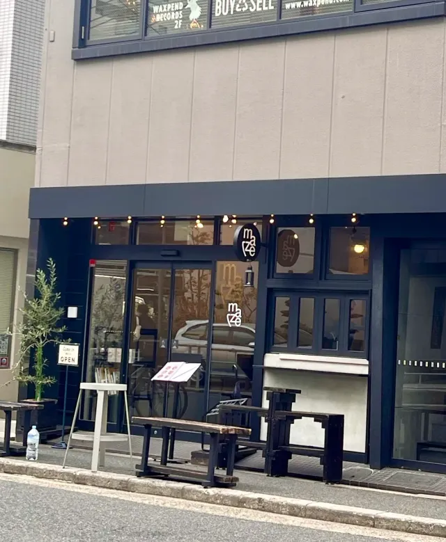 A dark front with a round sign, hanging bulbs, a potted olive tree and wooden benches on the street