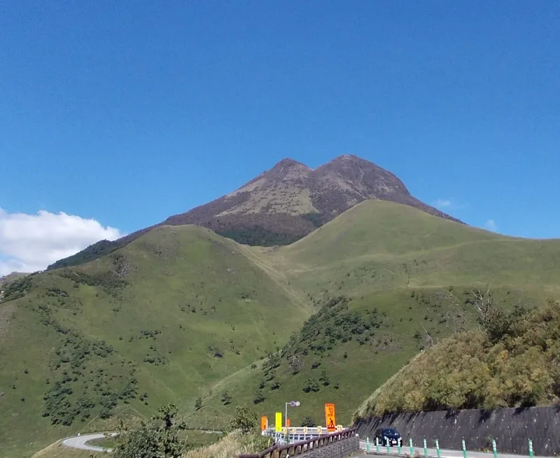 A bare twin peaked mountain above green grass slopes under a blue sky