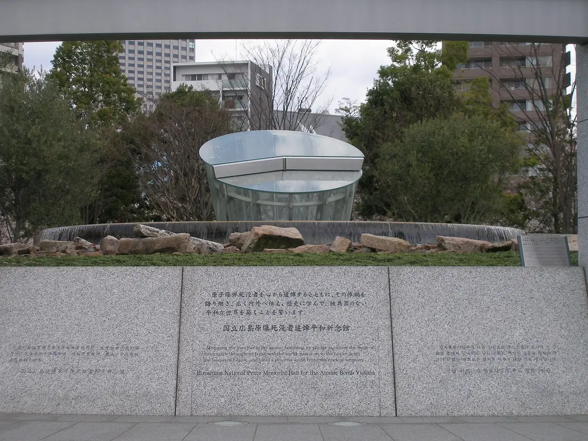 A granite wall with a dedication carved in Japanese and English, behind it the round glass structure of the memorial hall above a pool of water