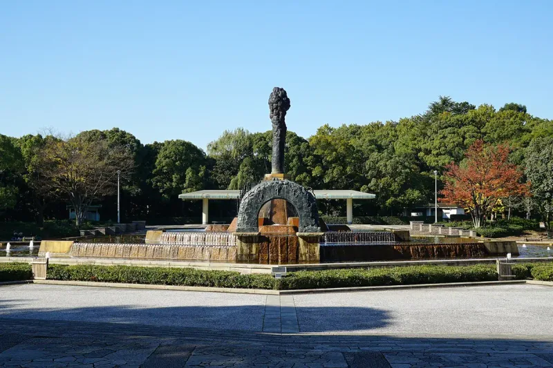 A plaza with a fountain and monument in a park, ringed by green and orange trees beside water