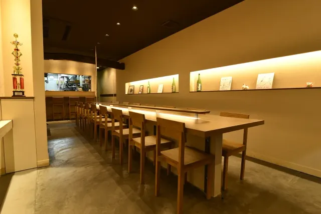 A long space with a long pale wooden table, chairs and lit niches with bottles