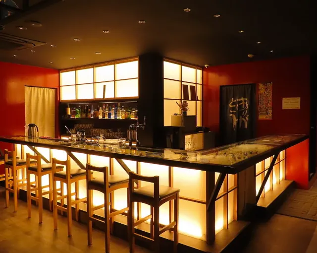 A counter lit from within with tall wooden stools, against red walls and shelves of bottles