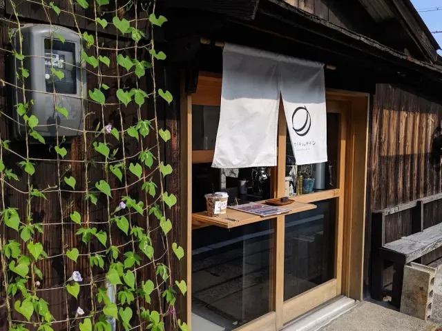 An old wooden house with a white cloth curtain, a small serving window and a climbing plant with flowers