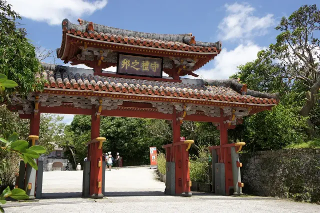 An ornate red wooden gate with a tiled roof, among green trees