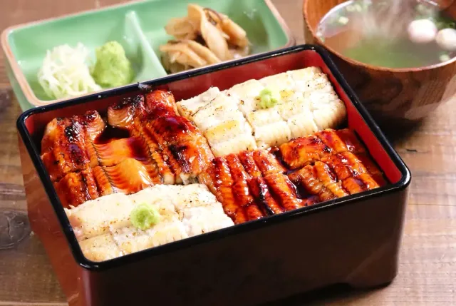 A lacquer box with slices of grilled eel over rice, beside soup and green pickles