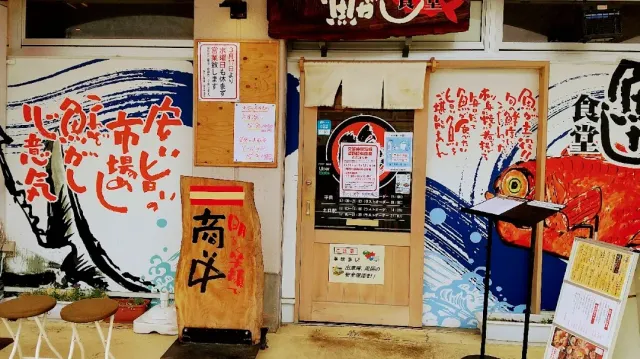 A colourful frontage with fish paintings and red Japanese script, a wooden sign and stools at the side